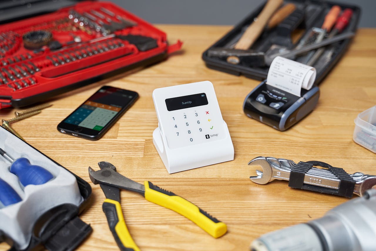 Red and black toolboxes next to smartphone and modern cash registers composing with pliers and wrenches on beige wooden table in bright room